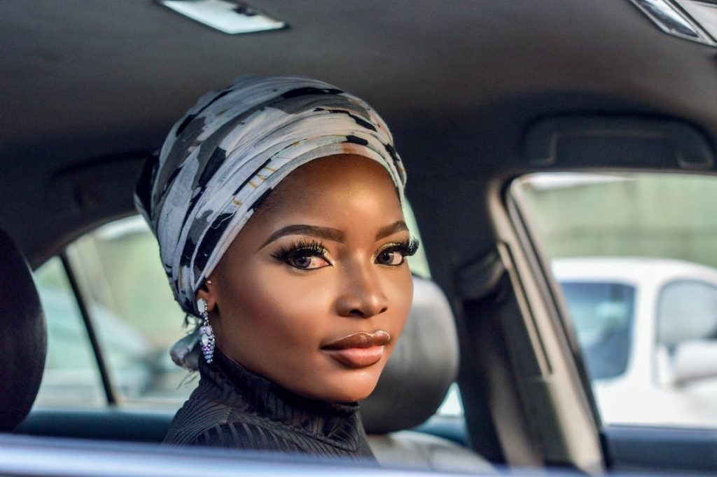 A woman with voluminous eyelashes is sitting inside a car while wearing a black and white floral headscarf